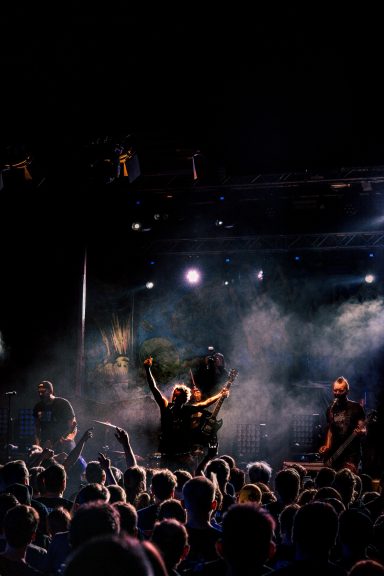 Dunkle Bühnenszene mit Band und Publikum, umgeben von Licht und Nebel.