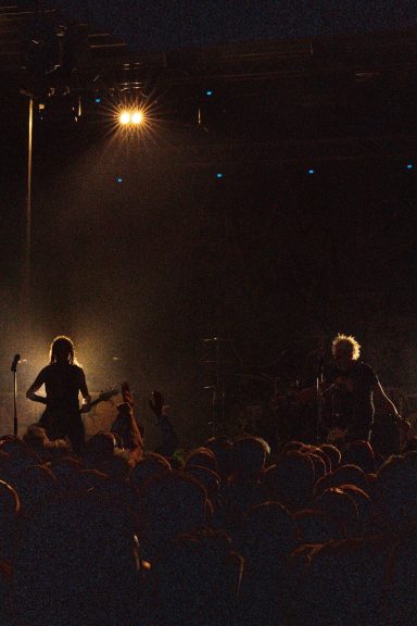 Dunkle Konzertszene mit Silhouetten von Musikern und Lichtstrahlen im Hintergrund.