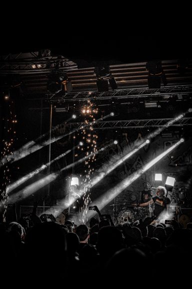 Dunkle Szene eines Konzerts mit Lichtstrahlen und Menschenmengen im Vordergrund.